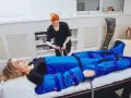 Kobieta leży na łóżku, podłączona do urządzenia do drenażu limfatycznego. Terapeutaka przygotowuje zabieg.