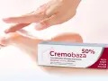 Dłoń delikatnie dotyka stopy, obok opakowanie kremu Cremobaza 50%. Produkt z mocznikiem, który pomaga zwalczać zrogowacenia.