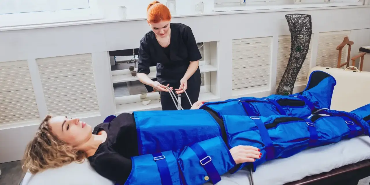 Kobieta leży na łóżku, podłączona do urządzenia do drenażu limfatycznego. Terapeutaka przygotowuje zabieg.