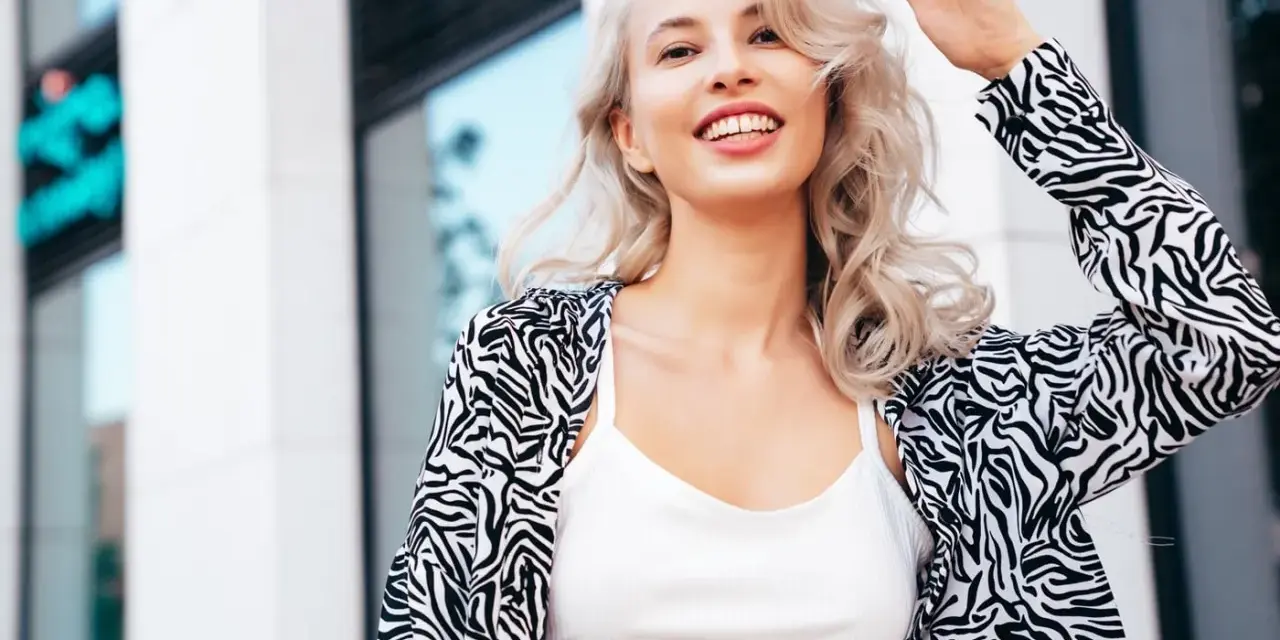 Uśmiechnięta blondynka z falowanymi włosami, efekt farbowania włosów na blond. Wzorzysta marynarka i biały top.
