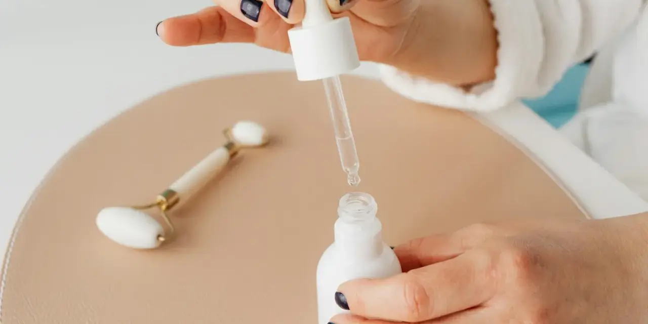 Dłoń z ciemnym lakierem do paznokci aplikuje serum z pipety do białej buteleczki. Obok leży jadeitowy roller. Zastanawiasz się, z czym łączyć niacynamid?