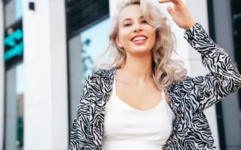 Uśmiechnięta blondynka z falowanymi włosami, efekt farbowania włosów na blond. Wzorzysta marynarka i biały top.