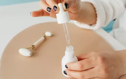 Dłoń z ciemnym lakierem do paznokci aplikuje serum z pipety do białej buteleczki. Obok leży jadeitowy roller. Zastanawiasz się, z czym łączyć niacynamid?