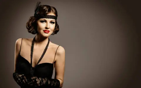Elegancka kobieta w stylu lat 20. z charakterystycznym makijażem, opaską na głowie i czarną suknią.
