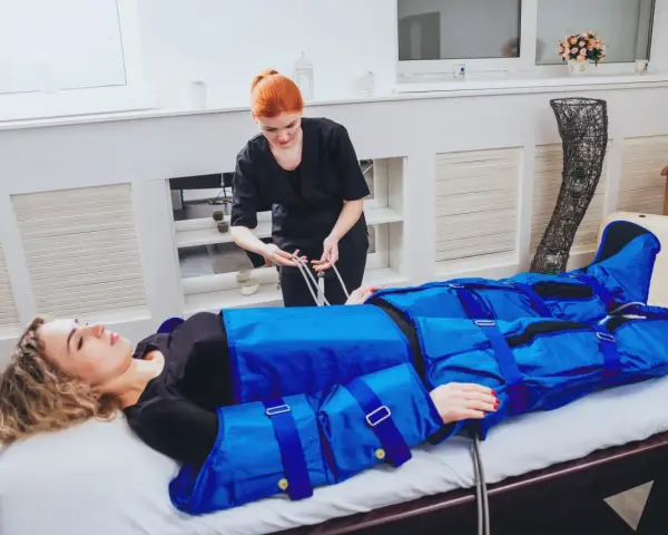 Kobieta leży na łóżku, podłączona do urządzenia do drenażu limfatycznego. Terapeutaka przygotowuje zabieg.