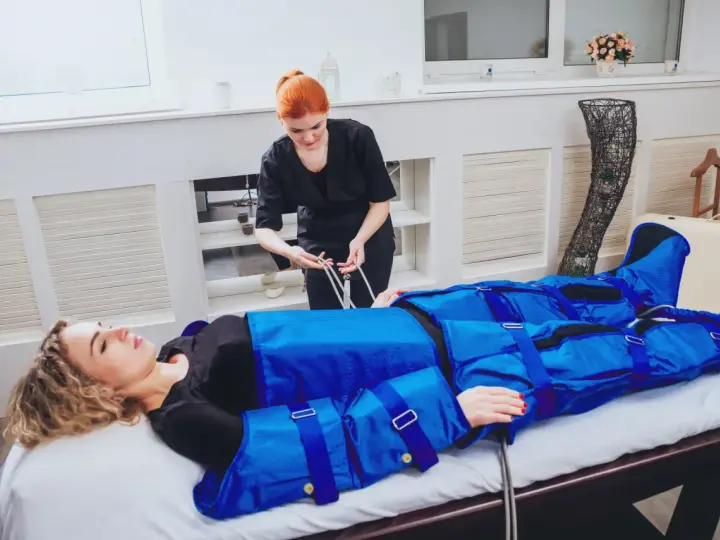 Kobieta leży na łóżku, podłączona do urządzenia do drenażu limfatycznego. Terapeutaka przygotowuje zabieg.