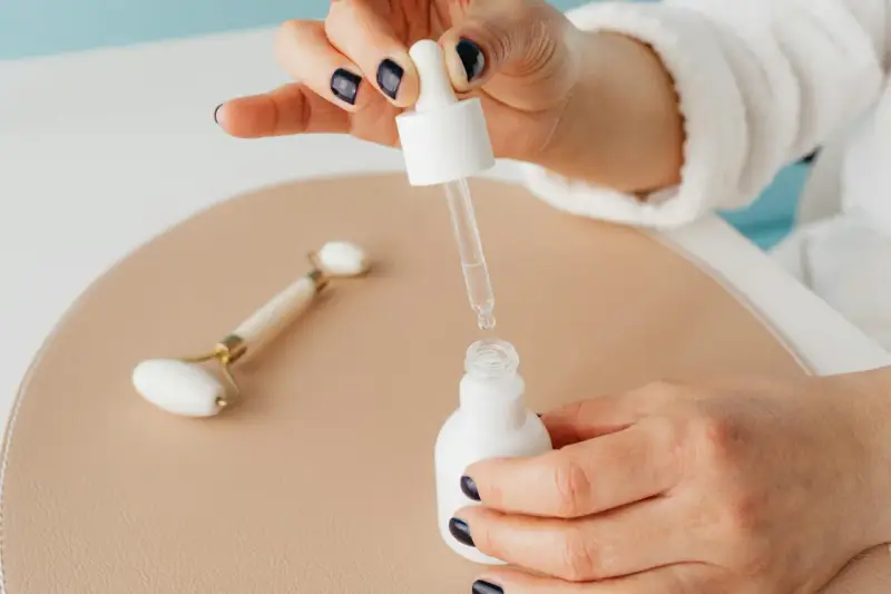 Dłoń z ciemnym lakierem do paznokci aplikuje serum z pipety do białej buteleczki. Obok leży jadeitowy roller. Zastanawiasz się, z czym łączyć niacynamid?